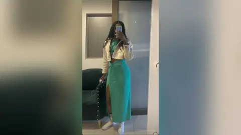 Young woman in a teal maxi skirt and white jacket taking a mirror selfie in a modern minimalist room.