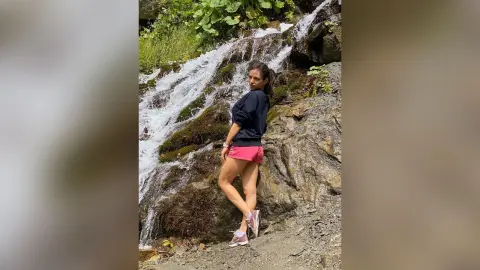 Young woman in black sweatshirt and pink shorts standing by a lush forest waterfall in a serene natural setting.