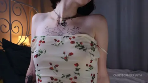 Young woman with chest tattoos wearing a floral cream tube top in a cozy bedroom setting.
