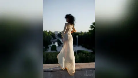 Elegant woman in a flowing white maxi dress standing on a balcony overlooking a lush green park at sunset.