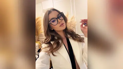 Stylish woman in a white blazer and glasses posing with a confident look in a bright indoor setting.