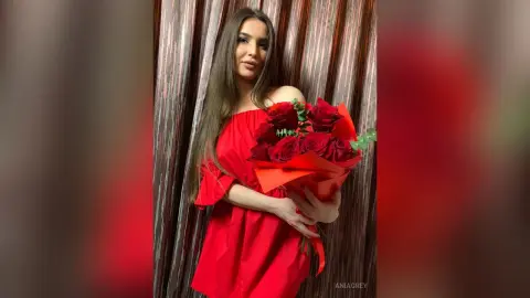 Beautiful woman in a red off-the-shoulder dress holding a large bouquet of red roses in a romantic setting.