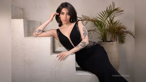 Elegant woman in a black evening gown leaning on white stone stairs next to a potted palm plant.