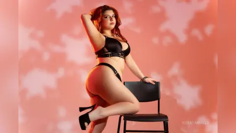 Curvy woman in black lingerie posing on a chair against a warm pink patterned background.