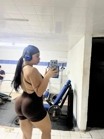 Athletic woman in brown gym shorts taking a mirror selfie in a bright fitness studio.