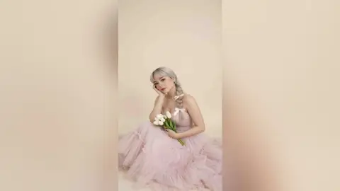Blonde woman in a dreamy pink tulle gown holding white tulips against a soft beige background.
