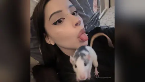 MarvellaRovella sticks her tongue out while posing with a small white and grey rat in her lap.
