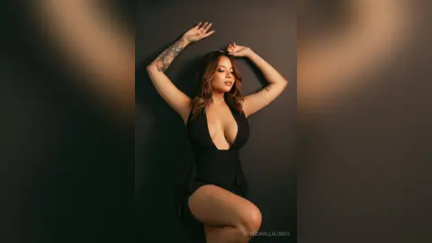 RebecaVillalobos poses with her arms raised against a dark wall, wearing a low-cut black romper or bodysuit.