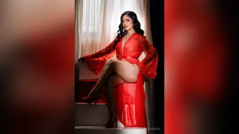 Elegant woman in a flowing red sheer robe sitting by a window in a romantic, moody setting.