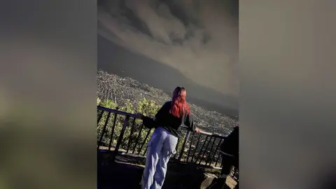 Woman in black top and light blue trousers overlooking a city view under a cloudy night sky.