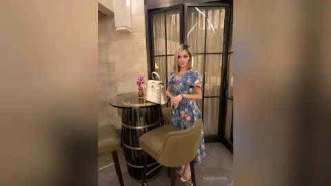 Elegant woman in a floral blue midi dress standing by a marble table in a modern, upscale interior setting.