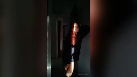 AmelieLautrec poses in a dark room, wearing a black shirt and showing her long red hair and tattooed leg.