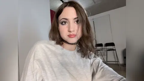AriaLush looks at the camera wearing a white t-shirt and a black lace choker with her wavy brown hair down.