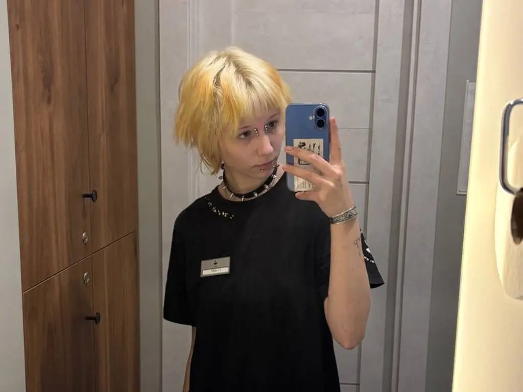 Blonde woman in a black t-shirt and choker taking a mirror selfie in a modern room.