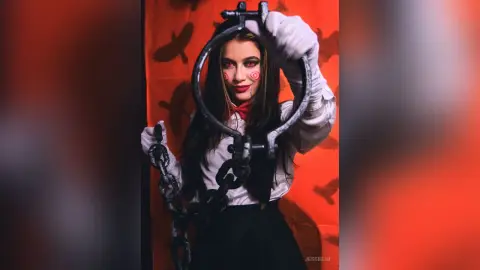 JessieLiu poses behind a large metal ring, wearing a white long-sleeved shirt and holding a heavy metal chain against a red background.