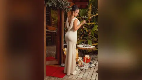 Elegant woman in a white backless evening gown holding a wine glass in a festive tropical garden setting.
