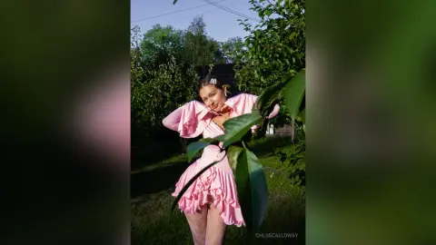 Smiling young woman in a pink ruffled dress posing in a lush green garden during golden hour.