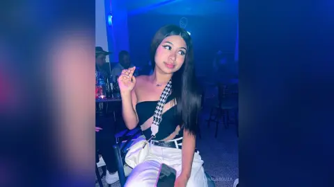 Young woman in a black crop top and white pants posing in a moody, blue-lit nightclub setting.