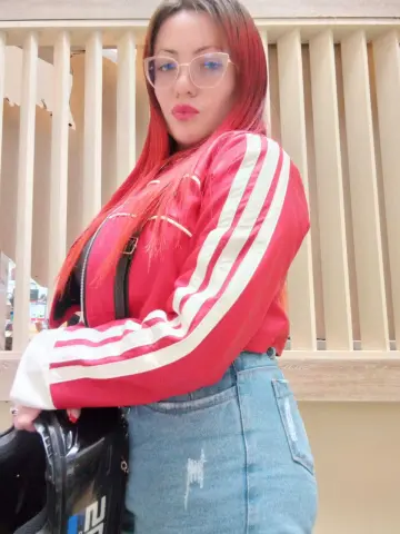 Red-haired woman in a red Adidas track jacket and denim jeans posing outdoors by a wooden fence.