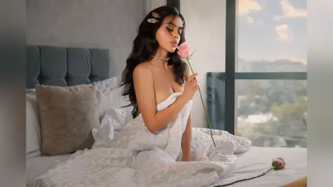 Dreamy portrait of a woman with long dark hair holding a pink rose while wrapped in white bedding in a bright bedroom.