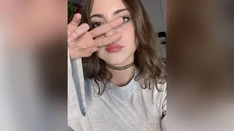 AriaLush poses with her hand over her eyes, wearing a grey long-sleeve shirt and a black lace choker.