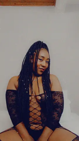 Smiling Black woman with long braids wearing a black lace cutout bodysuit in a bright indoor setting.