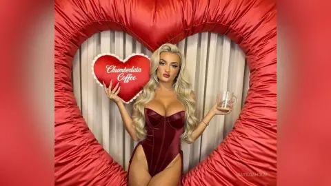Blonde woman in a red velvet bodysuit holding a Chamberlain Coffee heart pillow inside a giant red heart frame.