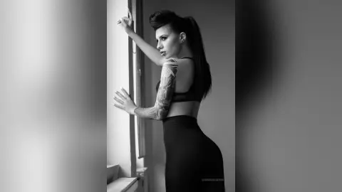 Moody black and white portrait of a tattooed woman in a black crop top looking out a window.