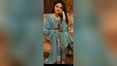 Smiling young woman in elegant light blue embroidered traditional ethnic outfit sitting on a patterned sofa.