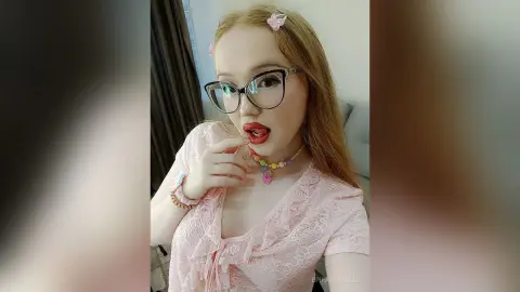 Young woman with glasses and pink lace top wearing colorful beaded jewelry in a bright indoor setting.