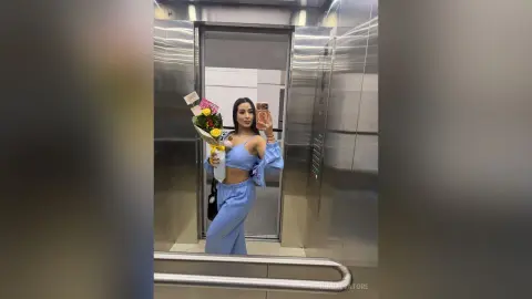Stylish woman in a blue two-piece outfit holding a flower bouquet in a modern elevator mirror selfie.