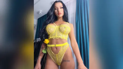 NicoleBella stands facing the camera wearing a yellow lace lingerie set with a yellow flower detail, showing her long black hair and tattoos.