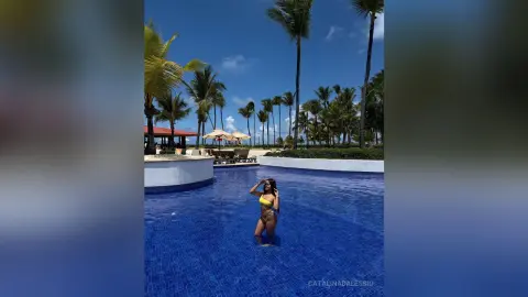 Woman in a yellow bikini relaxing in a tropical resort swimming pool surrounded by palm trees under a blue sky.