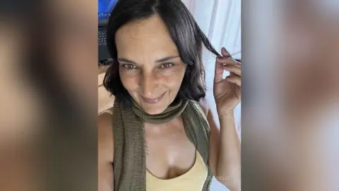 MichaelaBelle smiles while wearing a tan tank top and a dark olive scarf wrapped around her neck.
