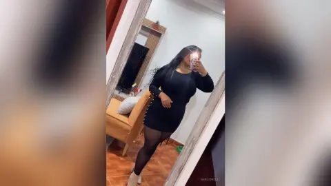 NiaCorelli poses in a black long-sleeve dress and black tights, taking a mirror selfie in a living room setting.