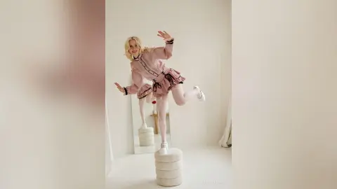 LynGoodhart poses playfully in a pink ruffled dress and tights while standing on a white pedestal in a bright room.