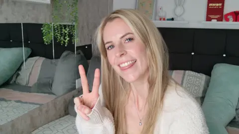 Smiling blonde woman in a cozy white sweater making a peace sign in a bright bedroom setting.