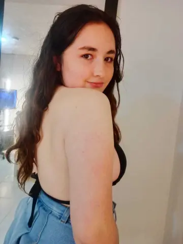 Young woman with long wavy hair wearing a black bikini top and denim shorts in a bright indoor setting.