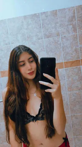 Young woman with long brown hair in a black lace bikini taking a mirror selfie in a tiled bathroom.