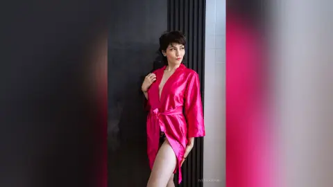 IveteDurand poses in a bright pink satin robe, showing off her legs and cleavage against a dark wall.