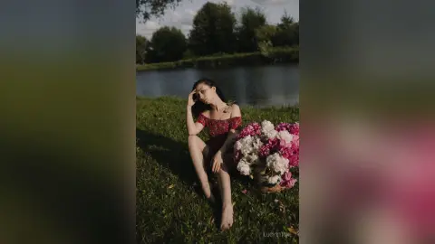 Dreamy portrait of a woman in a red lace dress sitting in a grassy field by a lake with pink peonies.