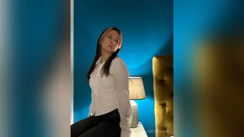 Young woman in a white blouse sitting in a moody blue bedroom setting.