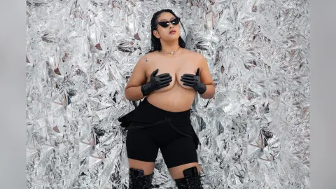 KimRoberth poses naked with black sunglasses and black gloves against a shiny silver tinsel background.