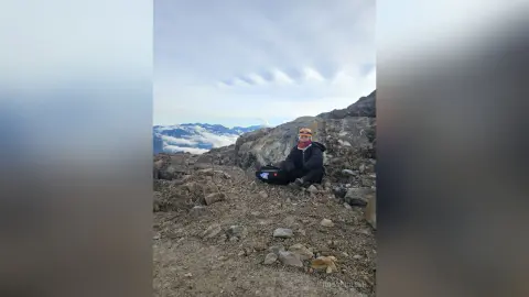 Hiker in a black jacket and orange beanie sitting on a rocky mountain peak overlooking a cloudy valley.