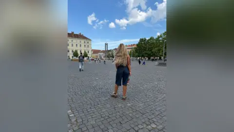 SelenaRusse stands in a cobblestone city square wearing dark denim shorts and a black top, looking out at the buildings under a blue sky.