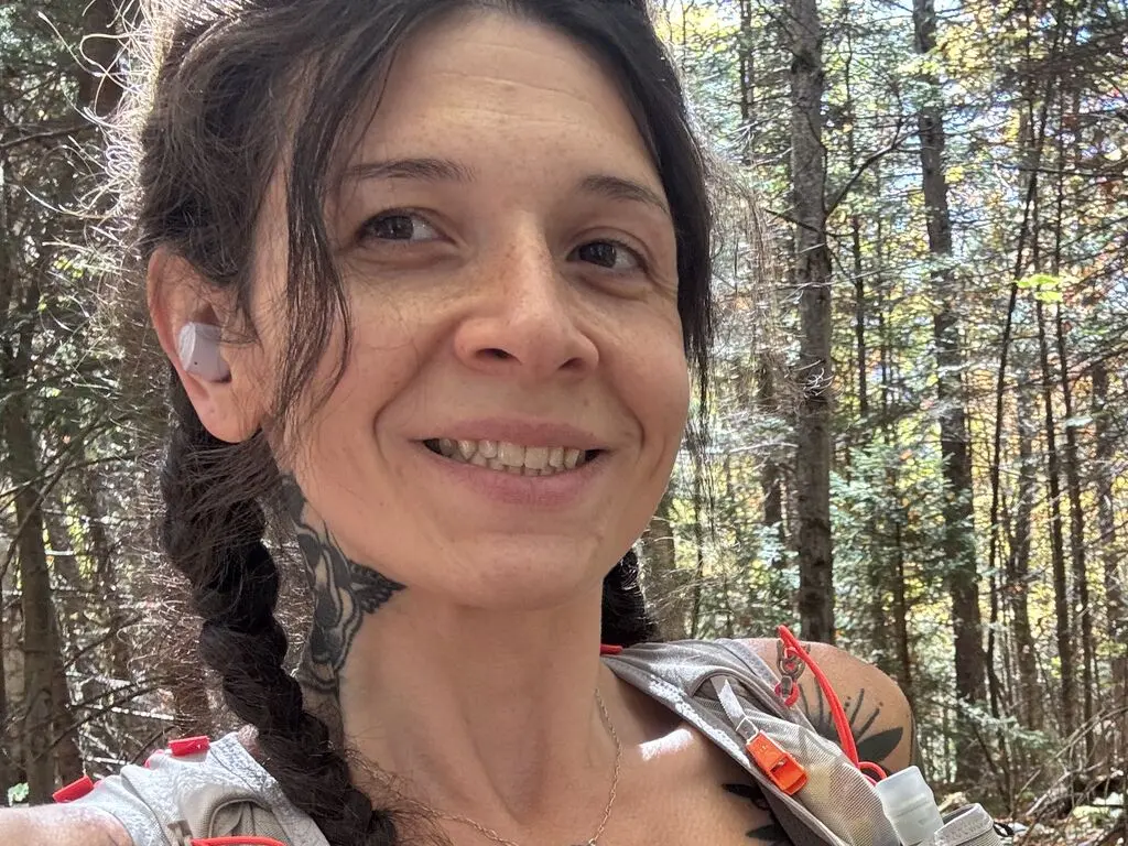 Smiling woman with braided hair and hiking gear enjoying a sunny day in a lush green forest.