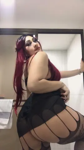 Woman with long red hair wearing a black lace bodysuit in a bright bathroom setting.