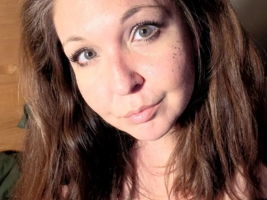 Close-up portrait of a smiling woman with long brown hair and green eyes in soft natural lighting.