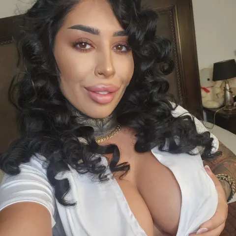 Smiling woman with long curly dark hair and neck tattoos wearing a white v-neck top in a cozy bedroom setting.
