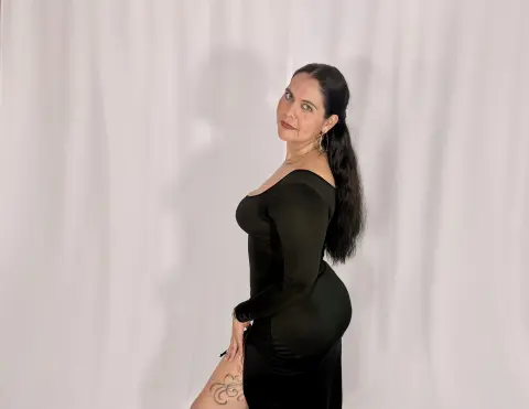Elegant woman in a sleek black long-sleeve dress posing against a white curtain background.